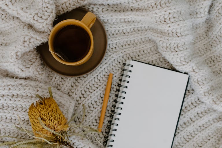 Warm flatlay with a cup of tea, a blank notebook, and pen on a knitted blanket.